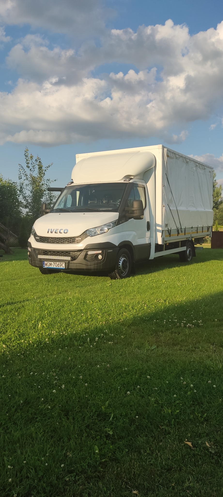 Biały samochód dostawczy Iveco z plandeką, zaparkowany na trawniku w słoneczny dzień.