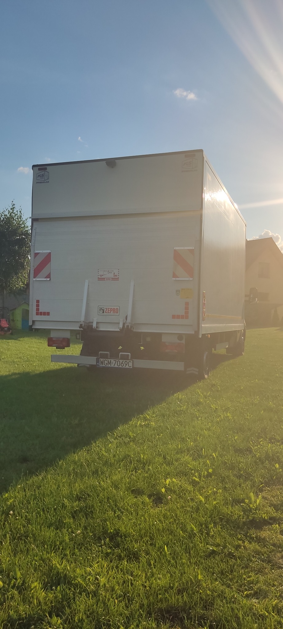 Tył białej ciężarówki z rampą załadowczą na zielonej trawie w słoneczny dzień, widoczne oznaczenia ostrzegawcze i tablica rejestracyjna WGM-7069C.