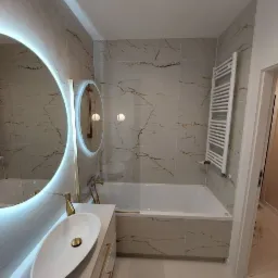 Nowoczesna łazienka z owalną umywalką, podświetlanym lustrem i płytkami imitującymi marmur z złotymi żyłami, grzejnik drabinkowy na ścianie.