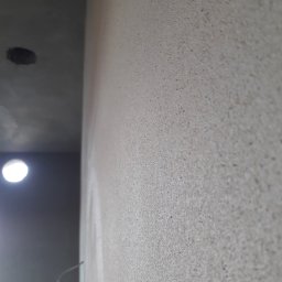 BARTO-TYNK - Ściana z szorstką, jasnoszarą fakturą, widoczny fragment sufitu z otworem na oświetlenie i wystający przewód elektryczny