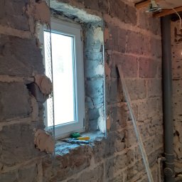 BARTO-TYNK - Montaż okna w surowej ścianie z kamienia, widoczne narzędzia na parapecie i metalowa siatka wzmacniająca wokół ramy.