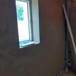 BARTO-TYNK - Świeżo otynkowana ściana wewnętrzna z widocznym oknem, na parapecie leży niebieski ręcznik, obok pionowa rura kanalizacyjna z łatą oparta o ścianę.