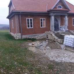 BARTO-TYNK - Dom w trakcie budowy z czerwonej cegły, widoczne białe okna, kolumny wejściowe, częściowo wykończona elewacja z kamienia oraz materiały budowlane na posesji.