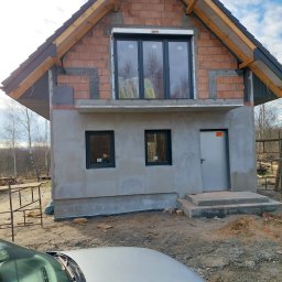 BARTO-TYNK - Surowa elewacja domu w trakcie budowy, z widocznymi cegłami, oknami z roletami i częściowo nałożonym szarym tynkiem. Na pierwszym planie fragment srebrnego samochodu.