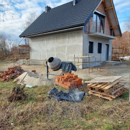 BARTO-TYNK - Surowy stan budynku w trakcie prac wykończeniowych z widoczną betonową elewacją, oknami, czarnym dachem z dachówki, rusztowaniami, stertą cegieł i betoniarką na zewnątrz w słoneczny dzień.