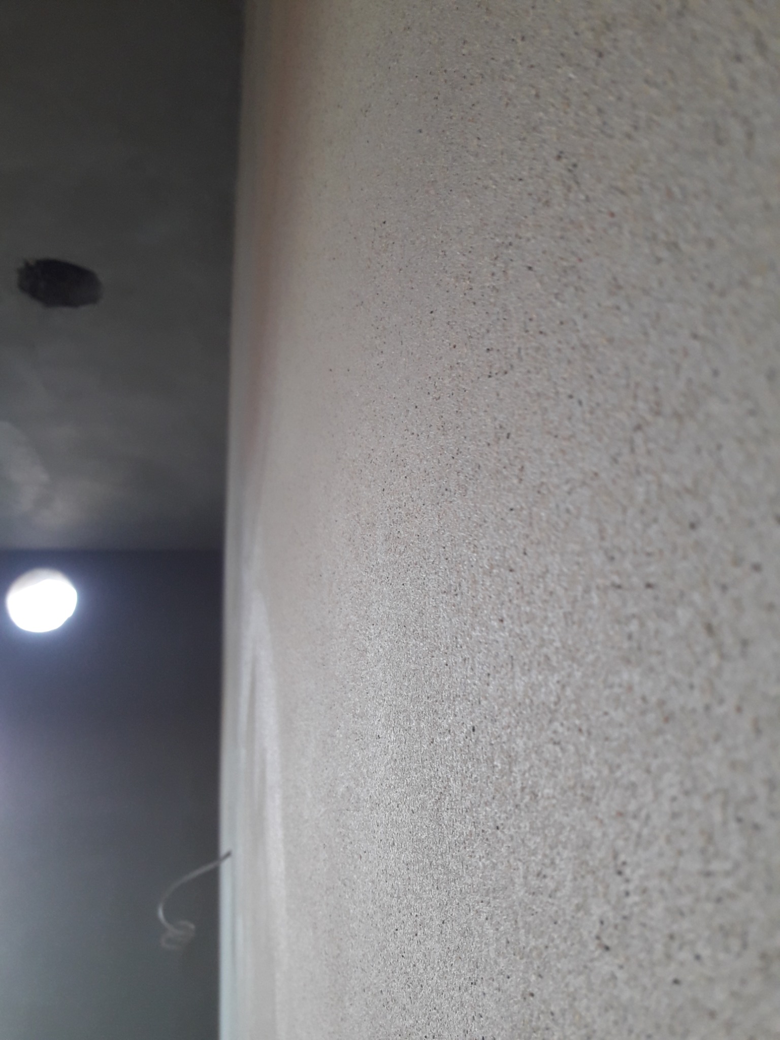 Ściana z szorstką, jasnoszarą fakturą, widoczny fragment sufitu z otworem na oświetlenie i wystający przewód elektryczny