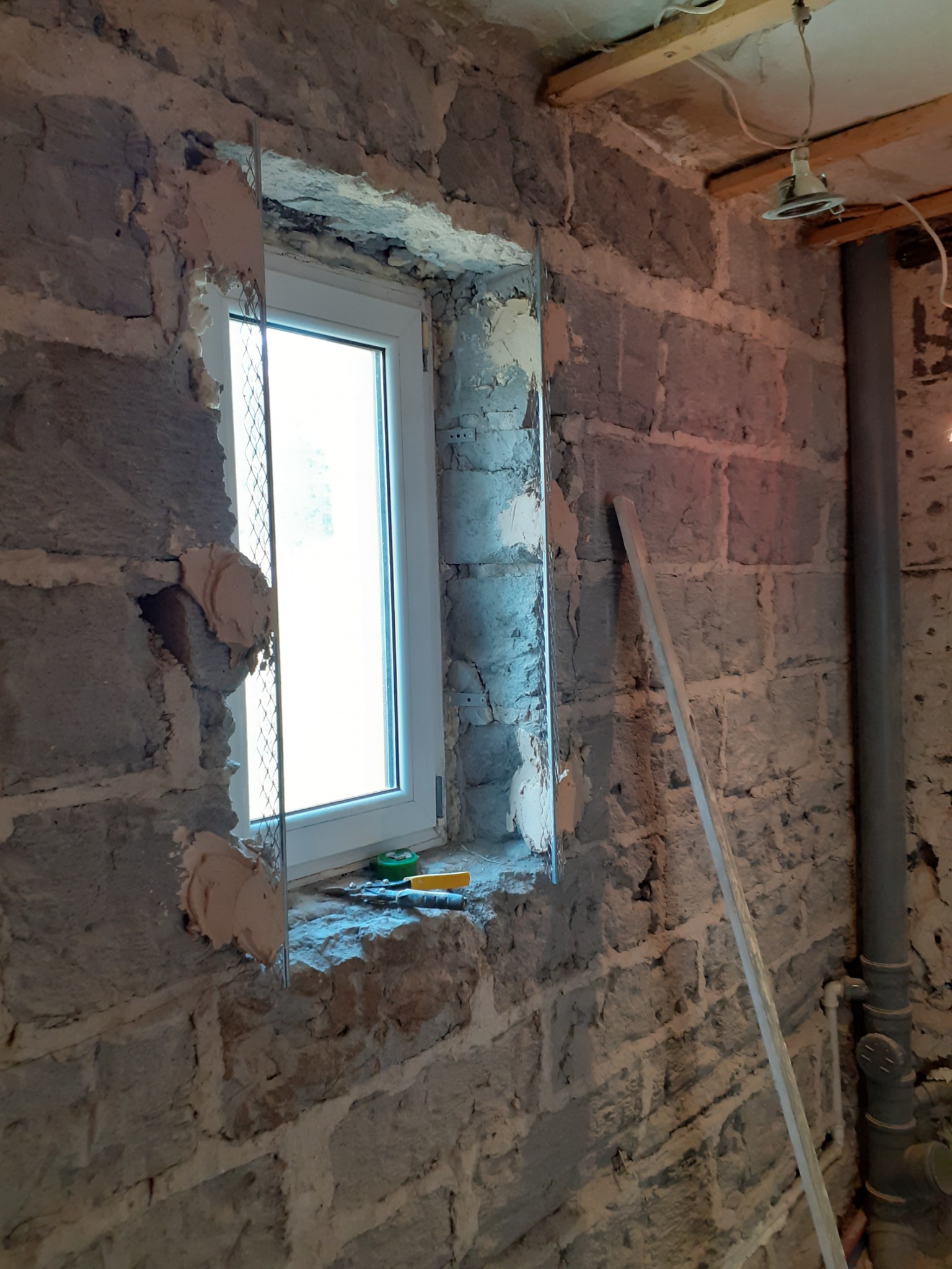 Montaż okna w surowej ścianie z kamienia, widoczne narzędzia na parapecie i metalowa siatka wzmacniająca wokół ramy.