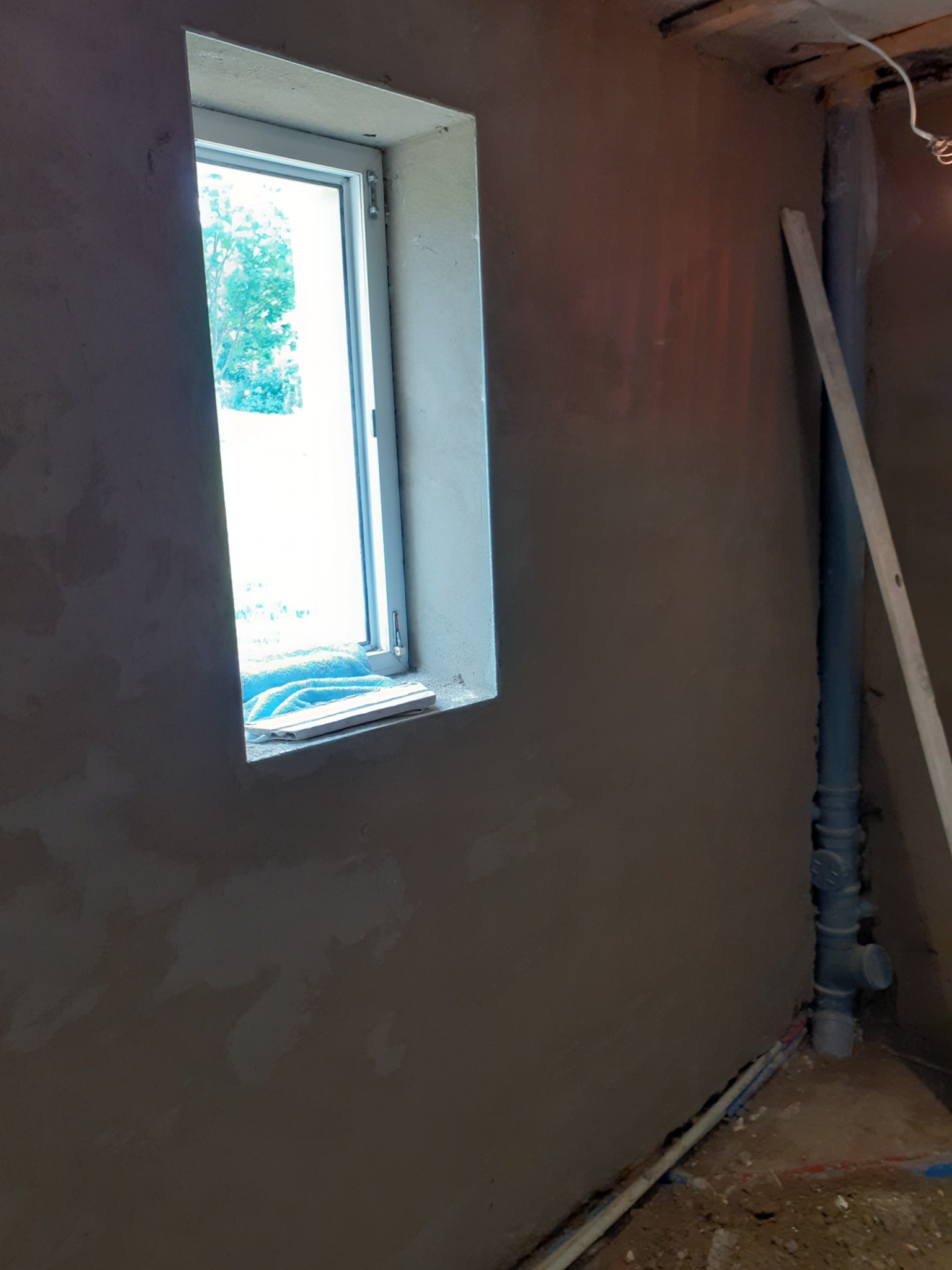 Świeżo otynkowana ściana wewnętrzna z widocznym oknem, na parapecie leży niebieski ręcznik, obok pionowa rura kanalizacyjna z łatą oparta o ścianę.