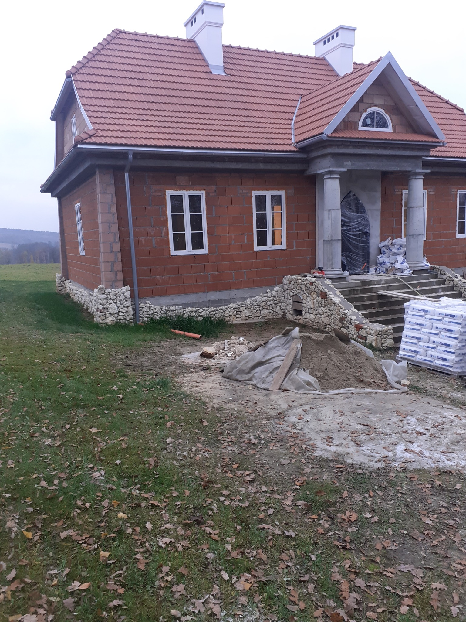 Dom w trakcie budowy z czerwonej cegły, widoczne białe okna, kolumny wejściowe, częściowo wykończona elewacja z kamienia oraz materiały budowlane na posesji.