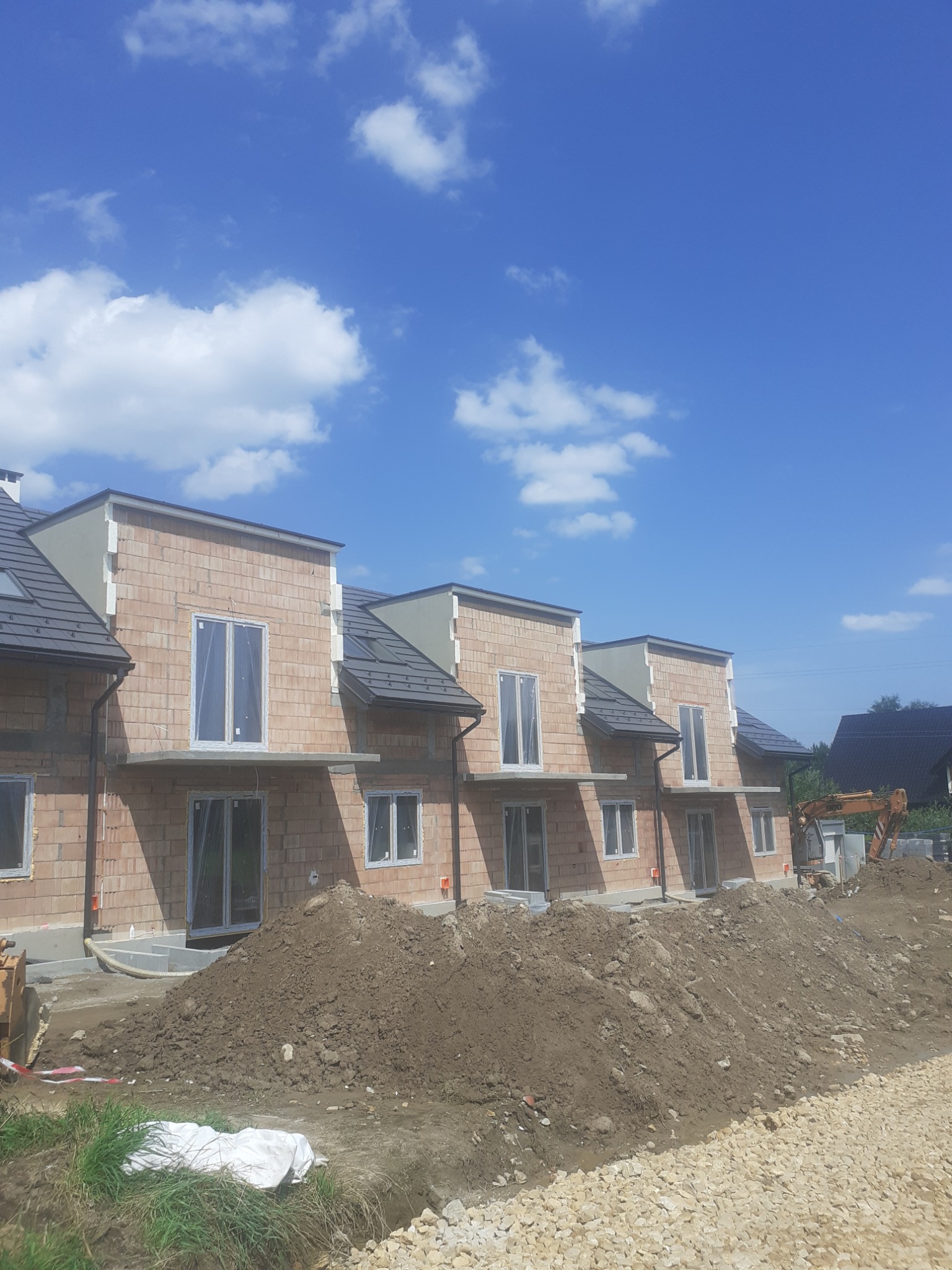 Budowa szeregu domów jednorodzinnych z cegły, widoczne okna zabezpieczone folią, na pierwszym planie hałda ziemi i gruzu, w tle koparka.