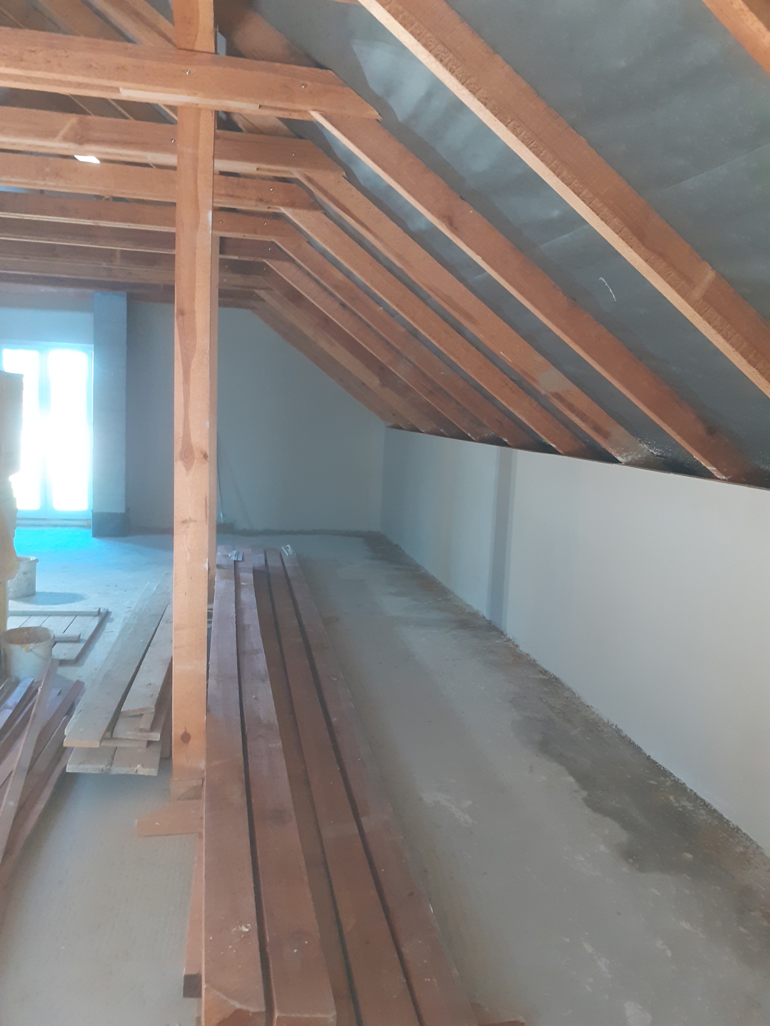 Konstrukcja drewniana poddasza z widocznymi belkami stropowymi i krokwiami, przygotowana do dalszych prac wykończeniowych, z ułożonymi deskami na podłodze i świeżo pomalowaną ścianą w tle.