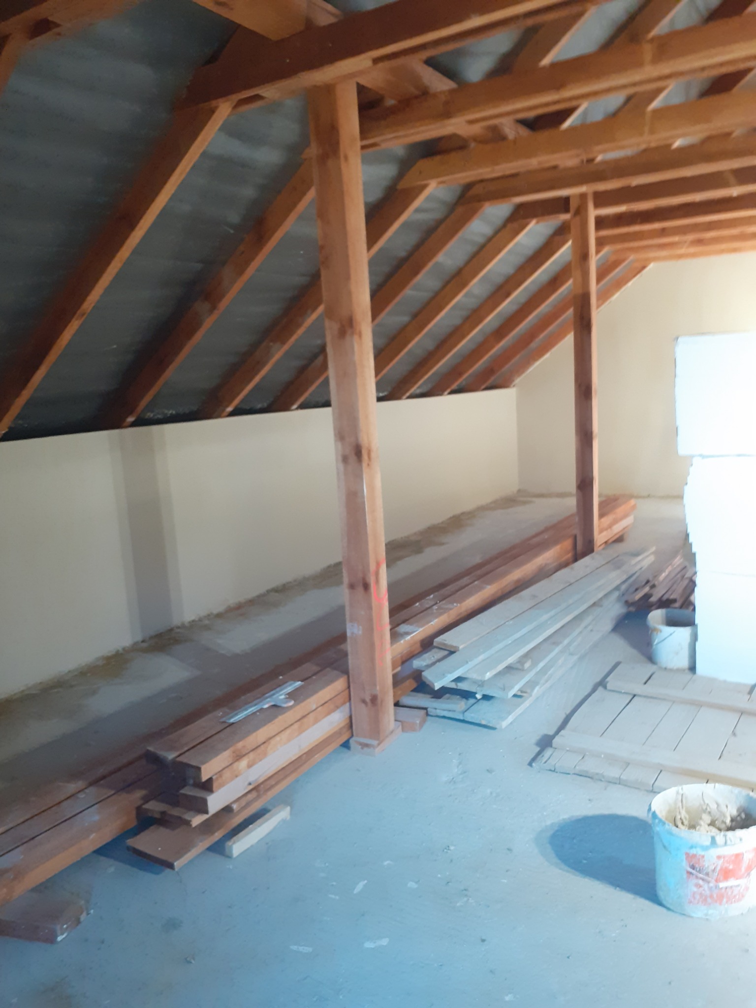 Poddasze w trakcie adaptacji: drewniana konstrukcja dachu, otynkowana ściana, przygotowane deski i płyty izolacyjne, widoczne ślady po pracach remontowych.