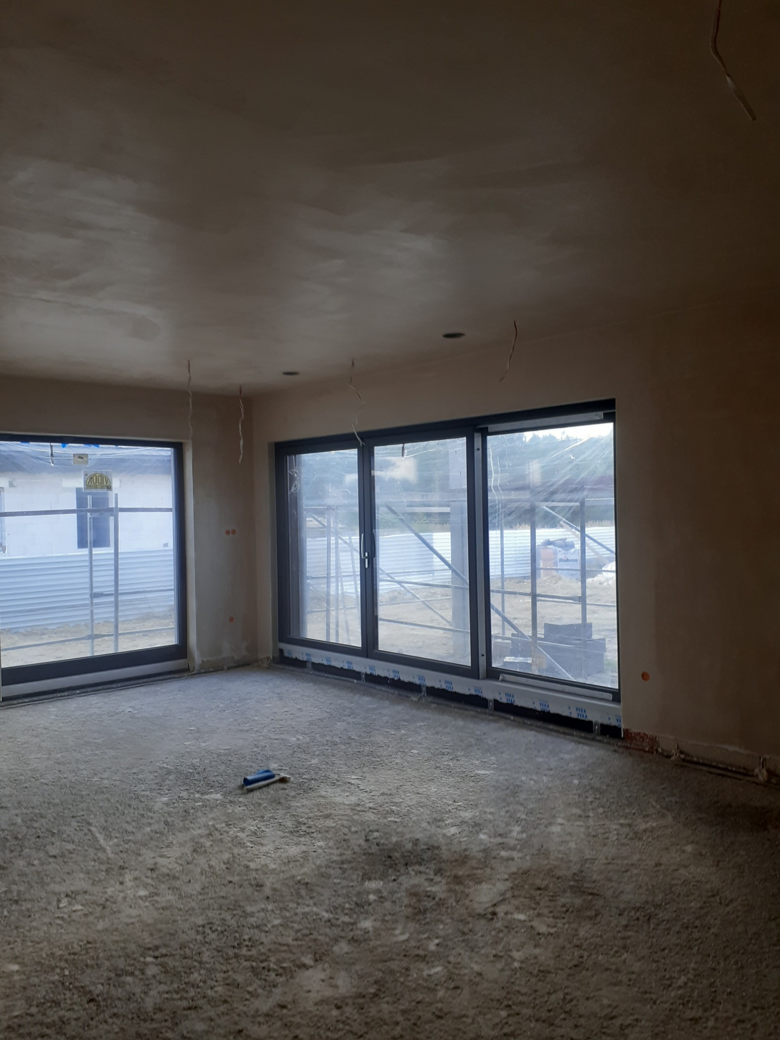 Surowe wnętrze nowo wybudowanego domu ze świeżo nałożonymi tynkami na ścianach i suficie, duże okna z widokiem na zewnątrz, zaprawa na podłodze i niebieski packa do gładzi leżąca na podłodze.