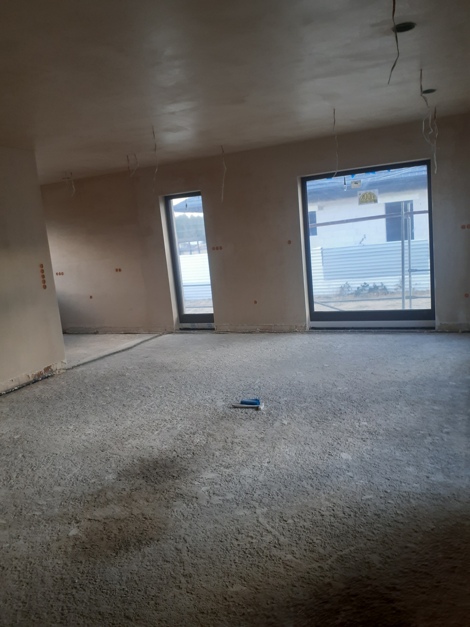 Surowe wnętrze w trakcie prac wykończeniowych: ściany pokryte świeżym tynkiem, widoczne otwory na gniazdka elektryczne i wystające kable z sufitu, surowa betonowa podłoga, okna z widokiem na budowę.