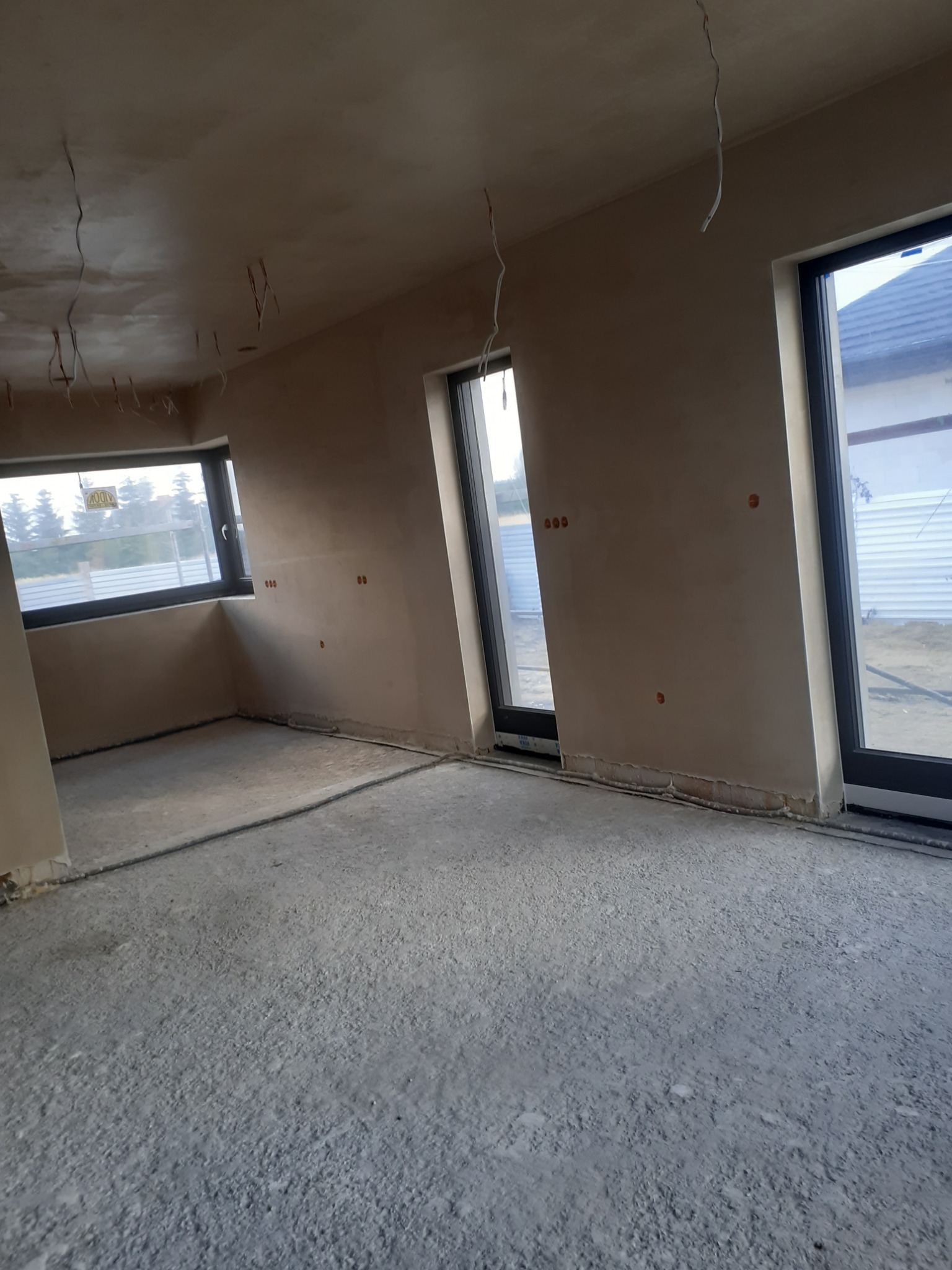 Surowe wnętrze po tynkowaniu maszynowym, widoczne wyprowadzone kable elektryczne, trzy okna z czarnymi ramami i szary betonowy podkład podłogowy.