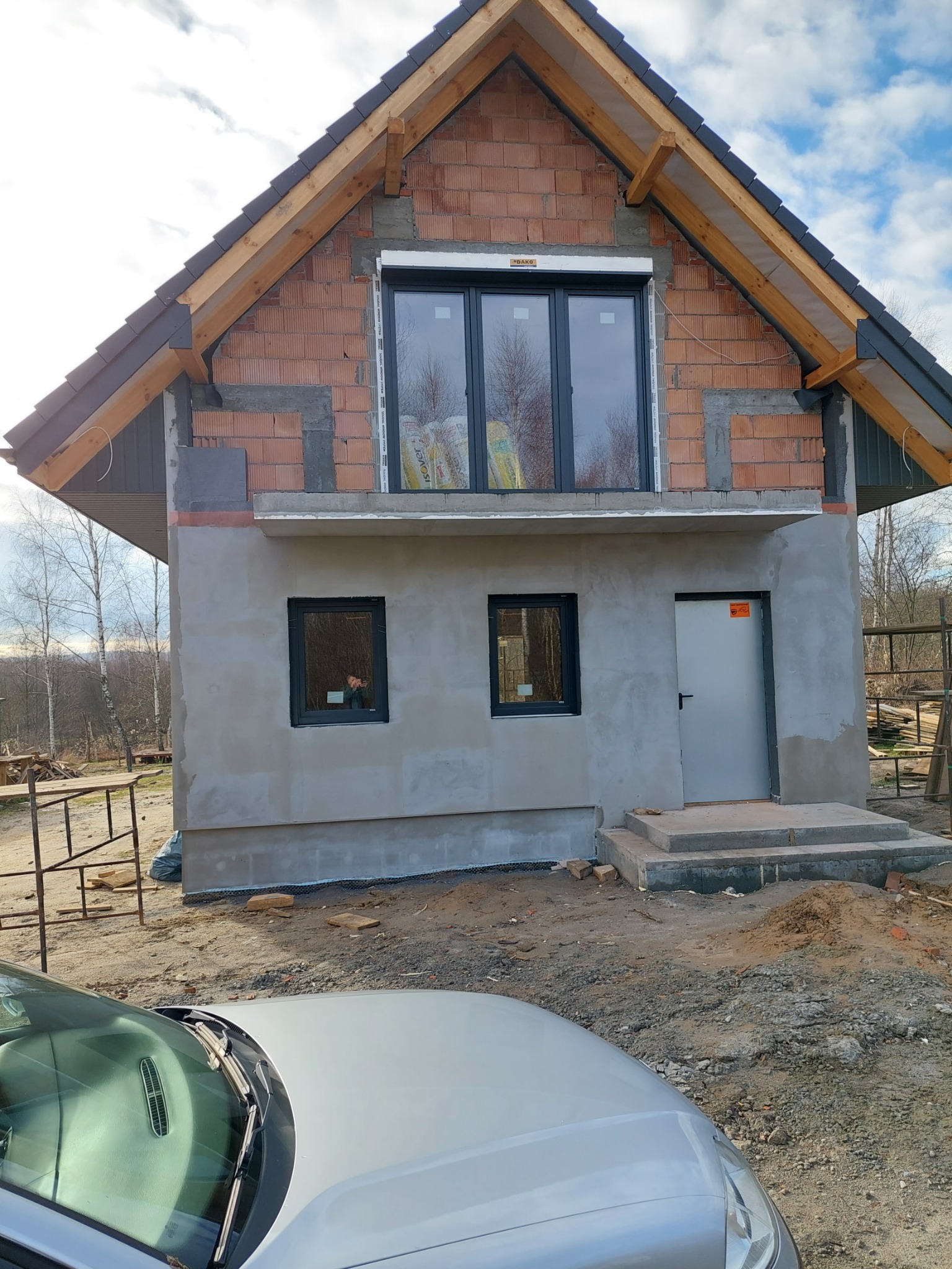 Surowa elewacja domu w trakcie budowy, z widocznymi cegłami, oknami z roletami i częściowo nałożonym szarym tynkiem. Na pierwszym planie fragment srebrnego samochodu.