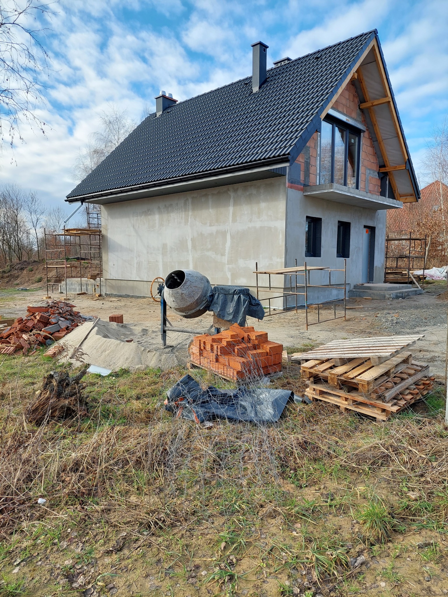 Surowy stan budynku w trakcie prac wykończeniowych z widoczną betonową elewacją, oknami, czarnym dachem z dachówki, rusztowaniami, stertą cegieł i betoniarką na zewnątrz w słoneczny dzień.