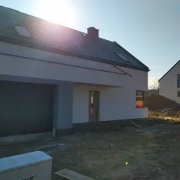 Nowoczesny dom jednorodzinny z szarą elewacją i ciemną dachówką, widoczny z zewnątrz w trakcie prac wykończeniowych, z niezagospodarowanym terenem wokół.