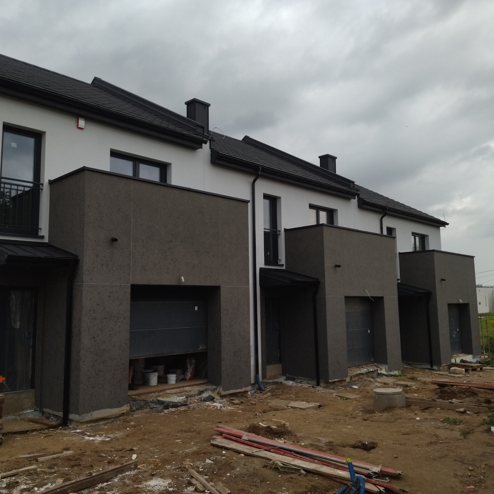 Szereg nowych domów w trakcie prac wykończeniowych elewacji, z widocznymi niedokończonymi garażami i materiałami budowlanymi na placu budowy.