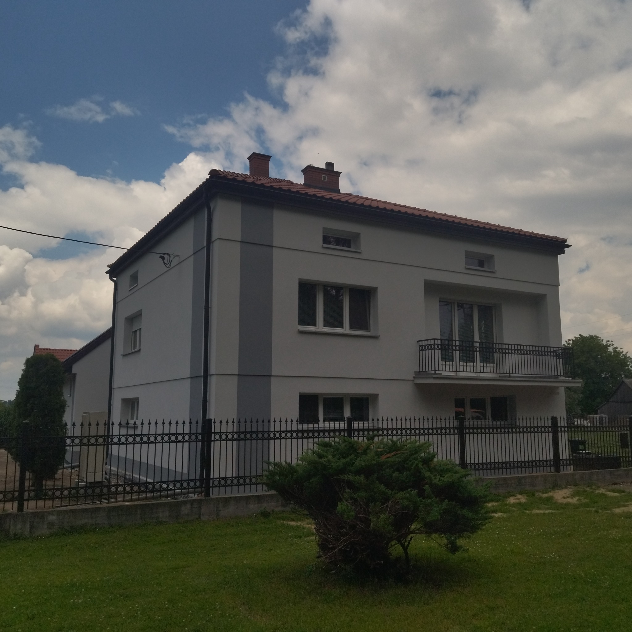 Dwu kondygnacyjny dom z jasnoszarą elewacją, ciemnoszarym pionowym pasem dekoracyjnym, balkonem z czarną balustradą i czerwoną dachówką, otoczony czarnym metalowym ogrodzeniem i zielonym trawnikiem.