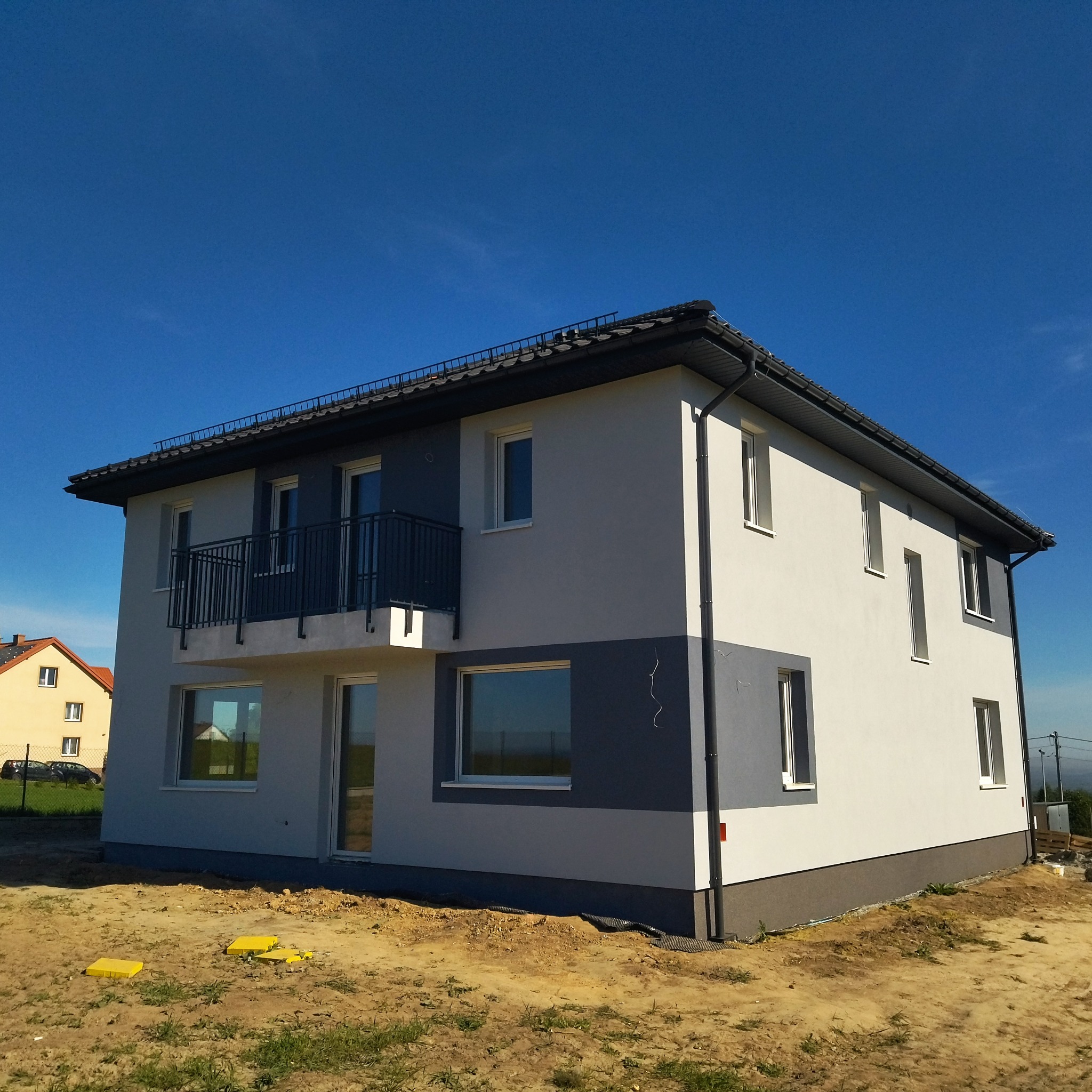Dwukondygnacyjny dom z balkonem, świeżo otynkowany w odcieniach szarości i bieli, na tle błękitnego nieba.