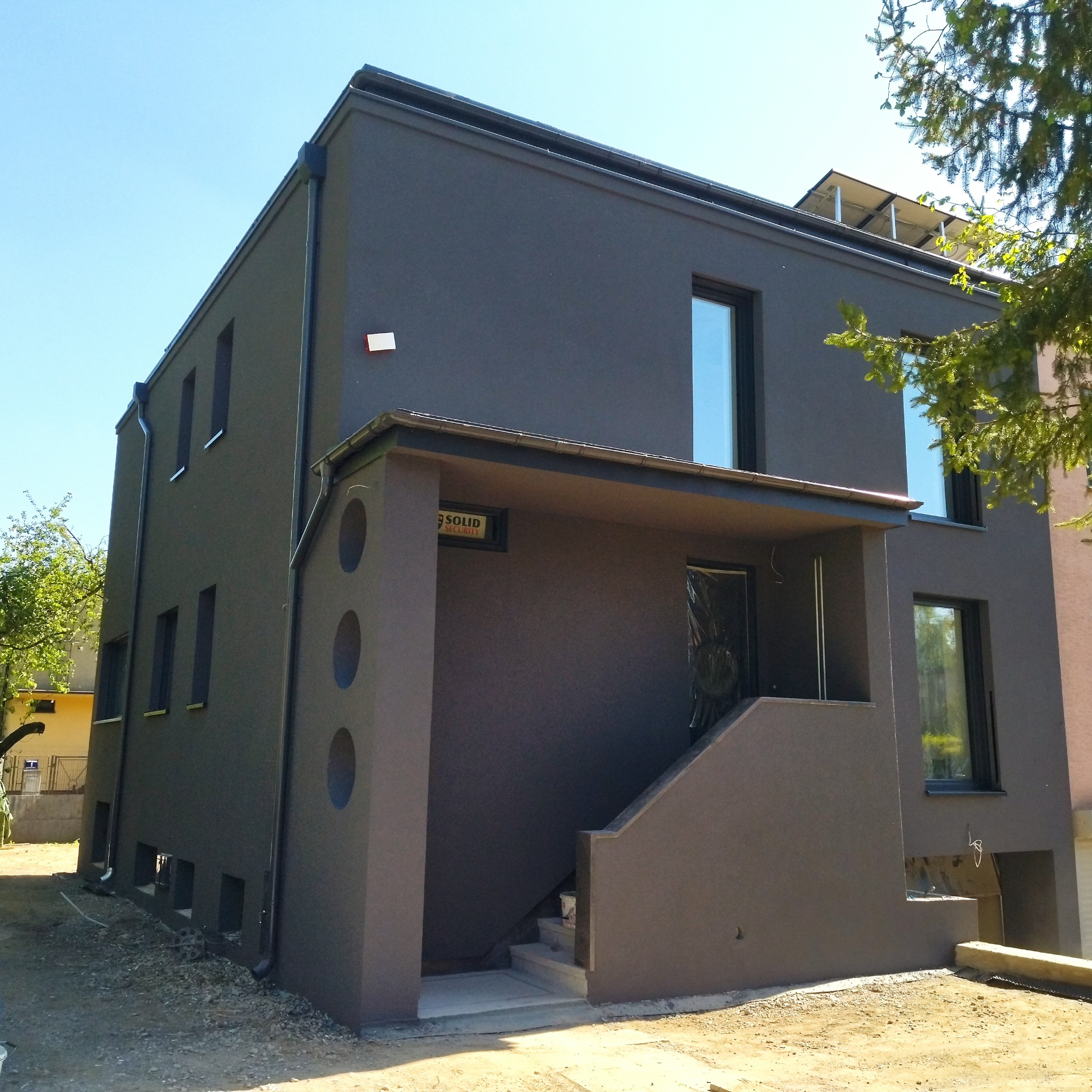 Nowoczesny, dwupiętrowy dom z ciemnoszarą elewacją, charakterystycznymi okrągłymi wgłębieniami przy wejściu i panelami słonecznymi na dachu.