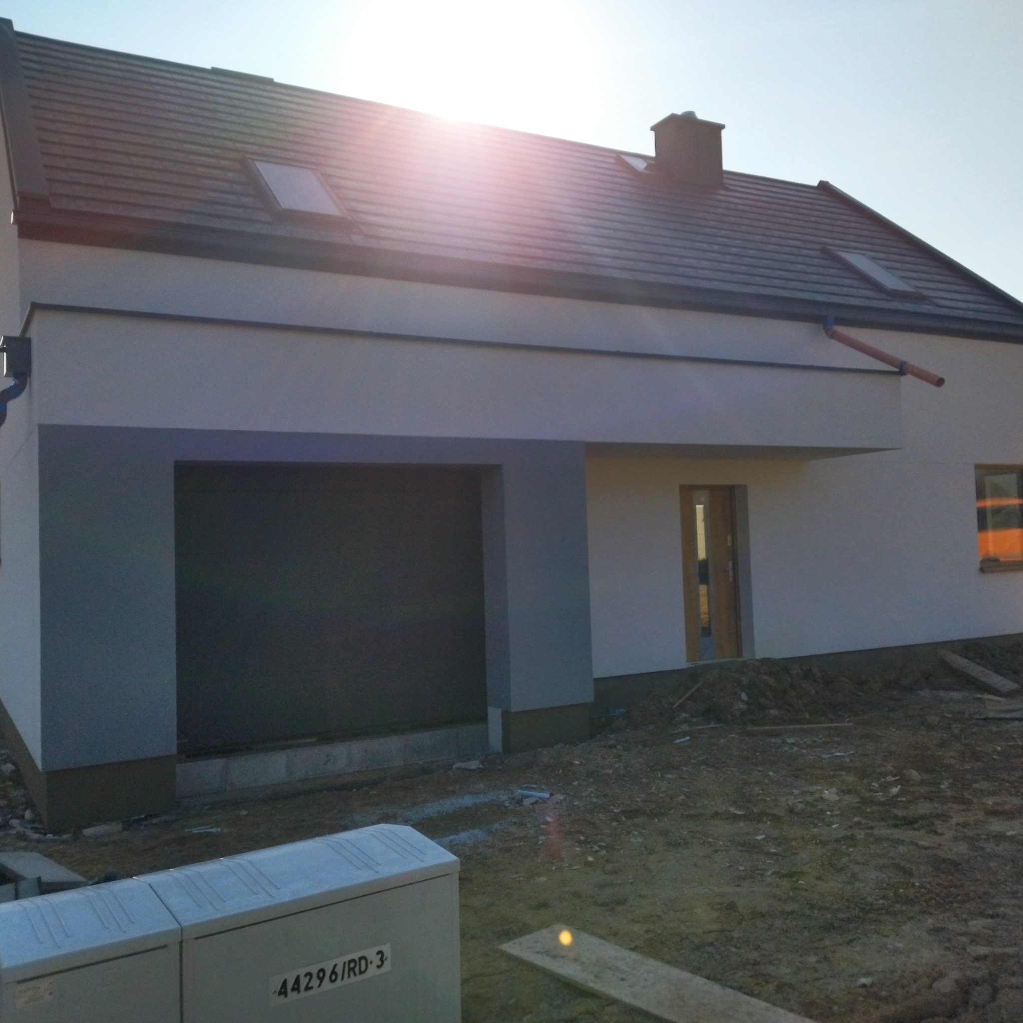 Nowoczesny dom jednorodzinny z szarą elewacją garażu i białym tynkiem, widoczne okna dachowe i rynna, teren wokół domu w trakcie prac wykończeniowych.