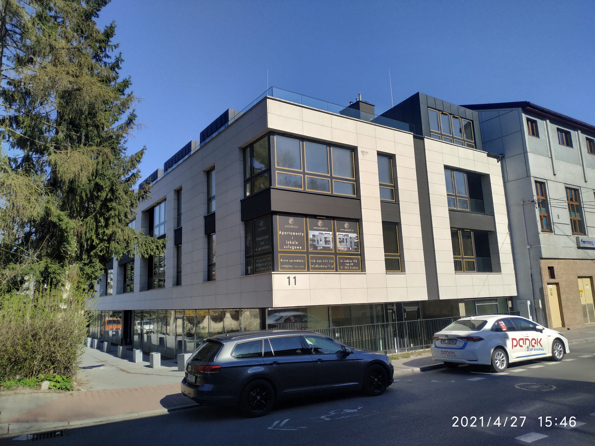 Nowoczesny budynek apartamentowy z elewacją w odcieniach beżu i szarości, z dużymi oknami i balkonami, widoczny z poziomu ulicy, z zaparkowanymi samochodami i drzewem po lewej stronie.