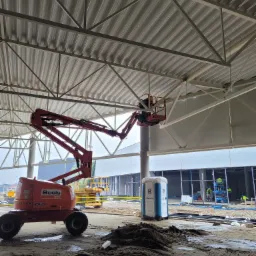 Montaż konstrukcji stalowej dachu hali z użyciem podnośnika koszowego Boels 450AJ, widoczne elementy konstrukcyjne dachu, pracownicy w oddali i tymczasowa toaleta na placu budowy.
