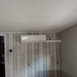 Mikola Maciej Kaczmarek - Biała jednostka wewnętrzna klimatyzacji marki Kaisai zamontowana na ścianie z paneli imitujących drewno, widoczny fragment okna z rośliną w metalowej doniczce.