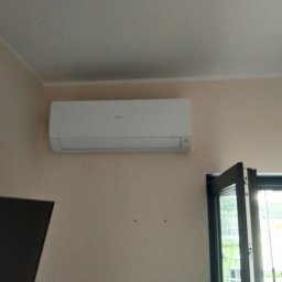 Mikola Maciej Kaczmarek - Biały klimatyzator ścienny zamontowany w jasnym pokoju, widoczne dwa otwory po poprzednim mocowaniu, okno uchylne w tle.