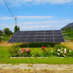 Panel słoneczny na metalowej konstrukcji, otoczony rabatą z kwitnącymi różami w odcieniach różu i bieli, na tle zielonego trawnika i błękitnego nieba z chmurami.