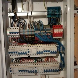 Mikola Maciej Kaczmarek - Rozdzielnica elektryczna w białej obudowie z widocznymi bezpiecznikami, przewodami w różnych kolorach, listwami zaciskowymi i ogranicznikiem przepięć, zamontowana przy ścianie.