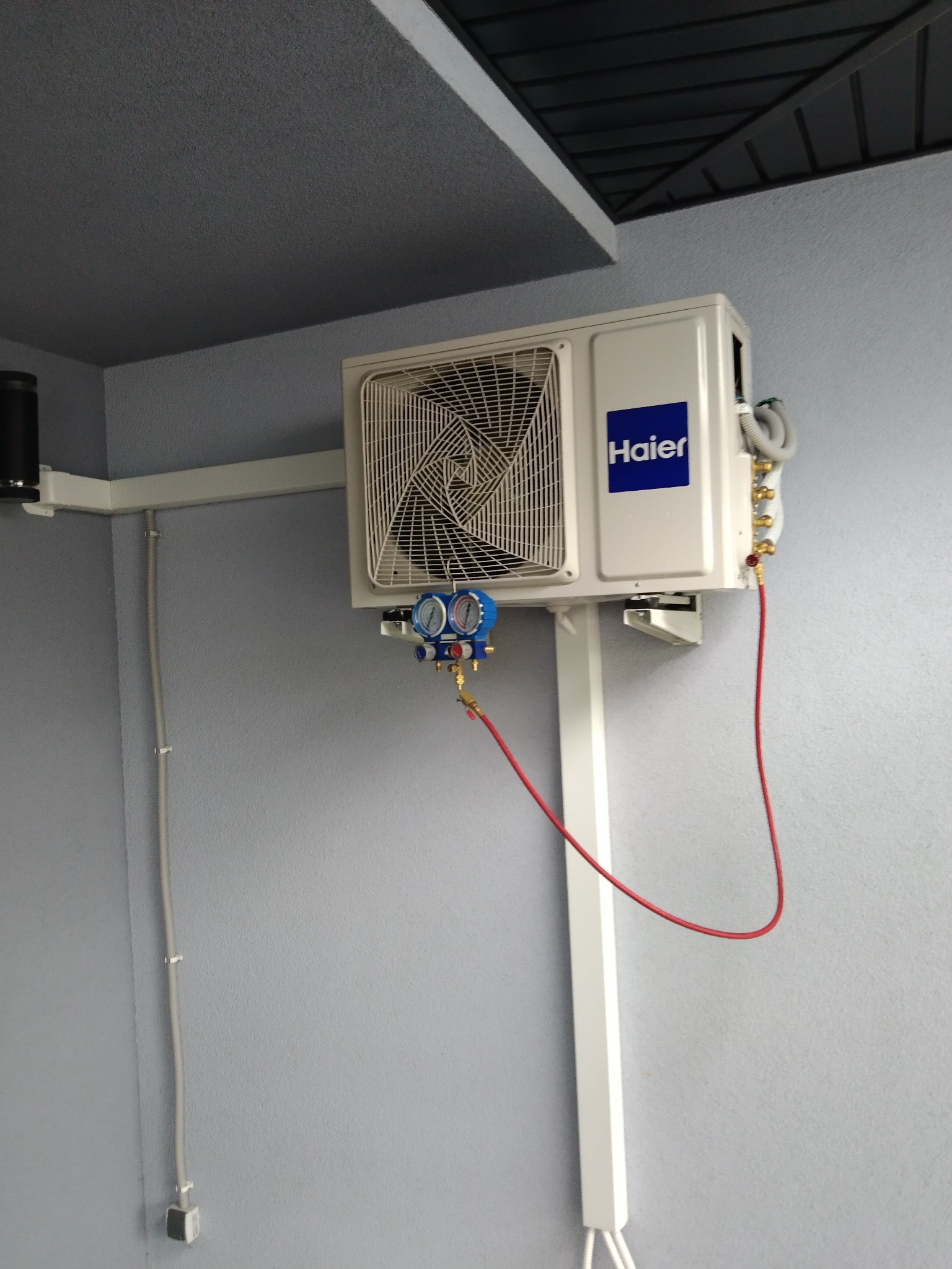 Biała jednostka zewnętrzna klimatyzacji Haier zamontowana na szarej ścianie, podłączona do manometrów za pomocą czerwonych węży, widoczne przewody elektryczne w listwach.