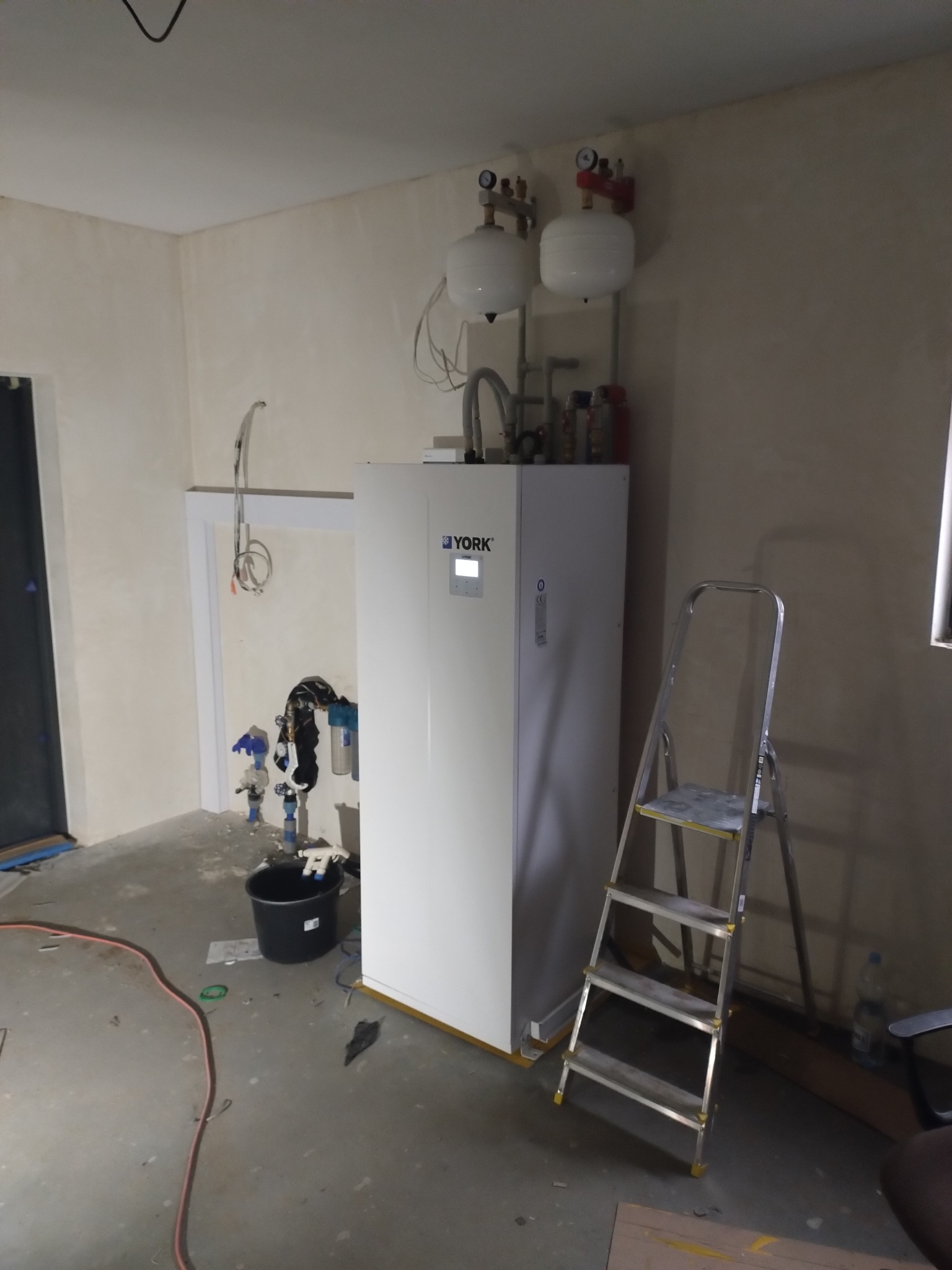 Biała pompa ciepła YORK w surowym pomieszczeniu podczas instalacji, widoczne zbiorniki wyrównawcze, drabina aluminiowa i elementy instalacji hydraulicznej w tle.