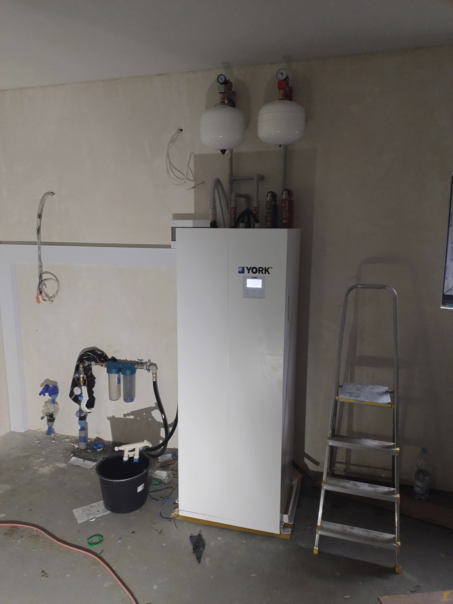 Biała pompa ciepła marki YORK w trakcie instalacji w surowym pomieszczeniu, widoczne podłączenia hydrauliczne, drabina i wiadro na podłodze.