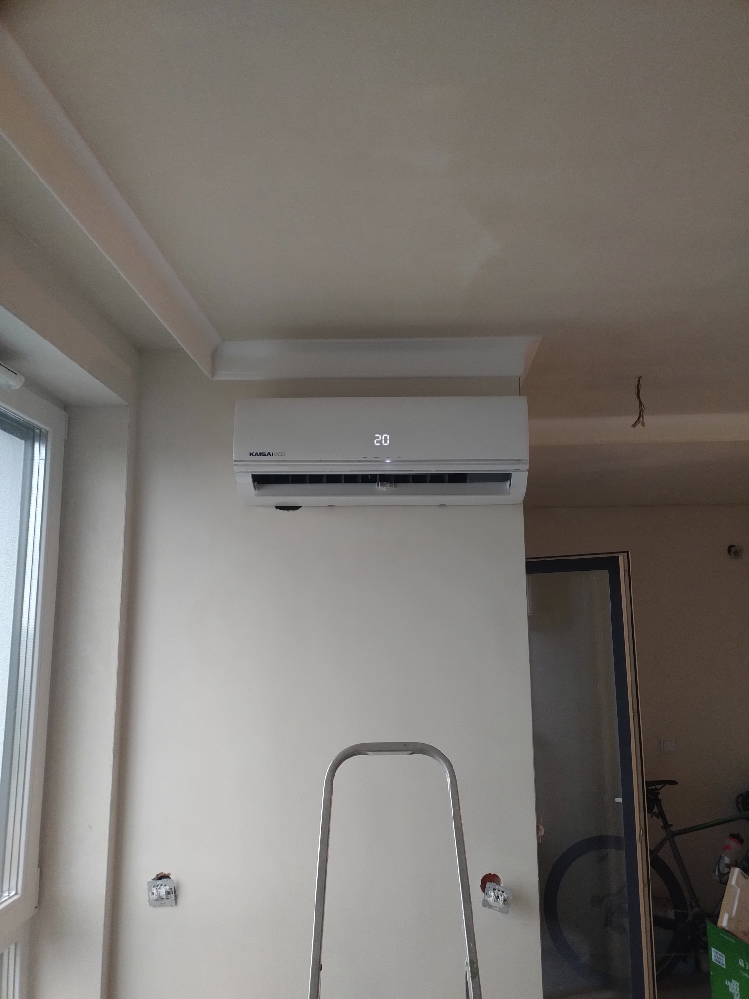 Biały klimatyzator Kaisaieco zamontowany na ścianie, widoczna drabina i wystające przewody elektryczne, sugerujące trwający montaż.