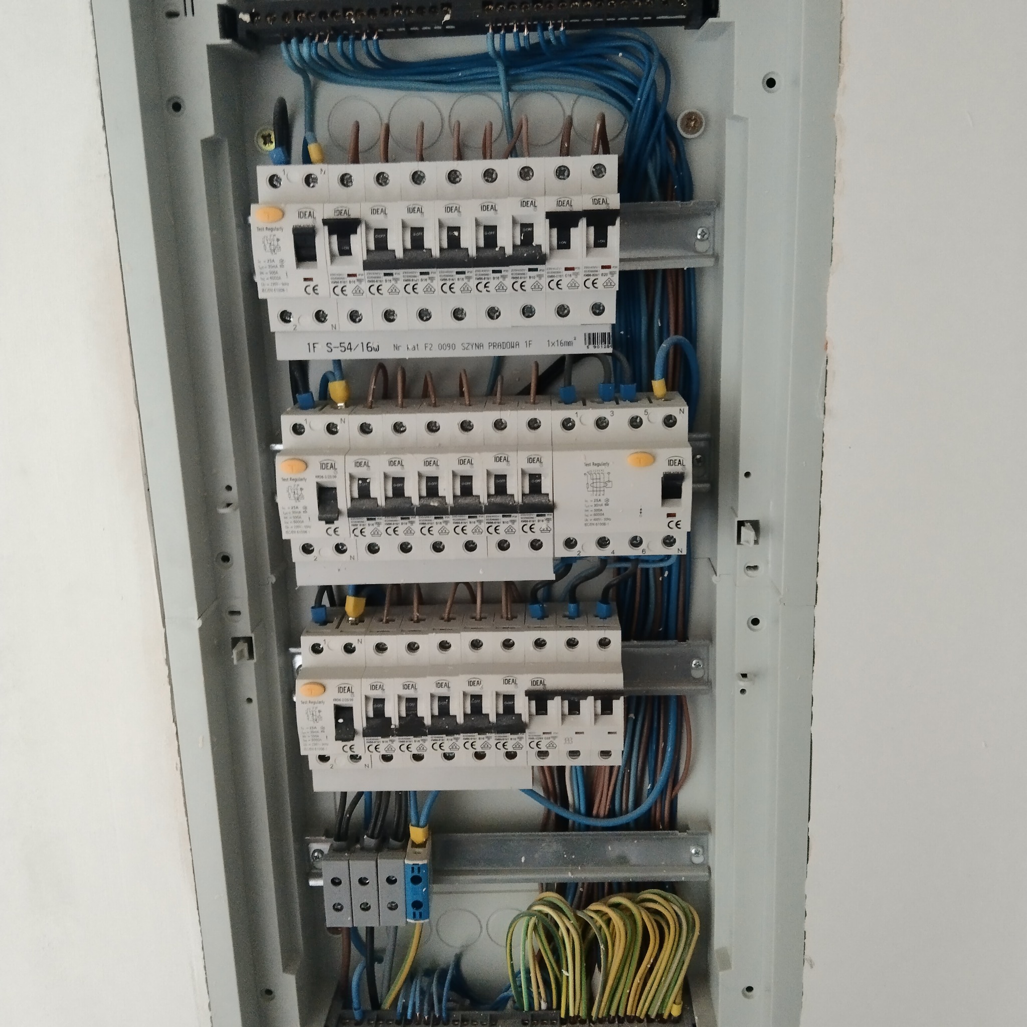 Rozdzielnica elektryczna z zamontowanymi bezpiecznikami IDEAL, widoczne kable w kolorach niebieskim, brązowym i żółto-zielonym, zamocowane na szynach wewnątrz obudowy, surowa ściana w tle.