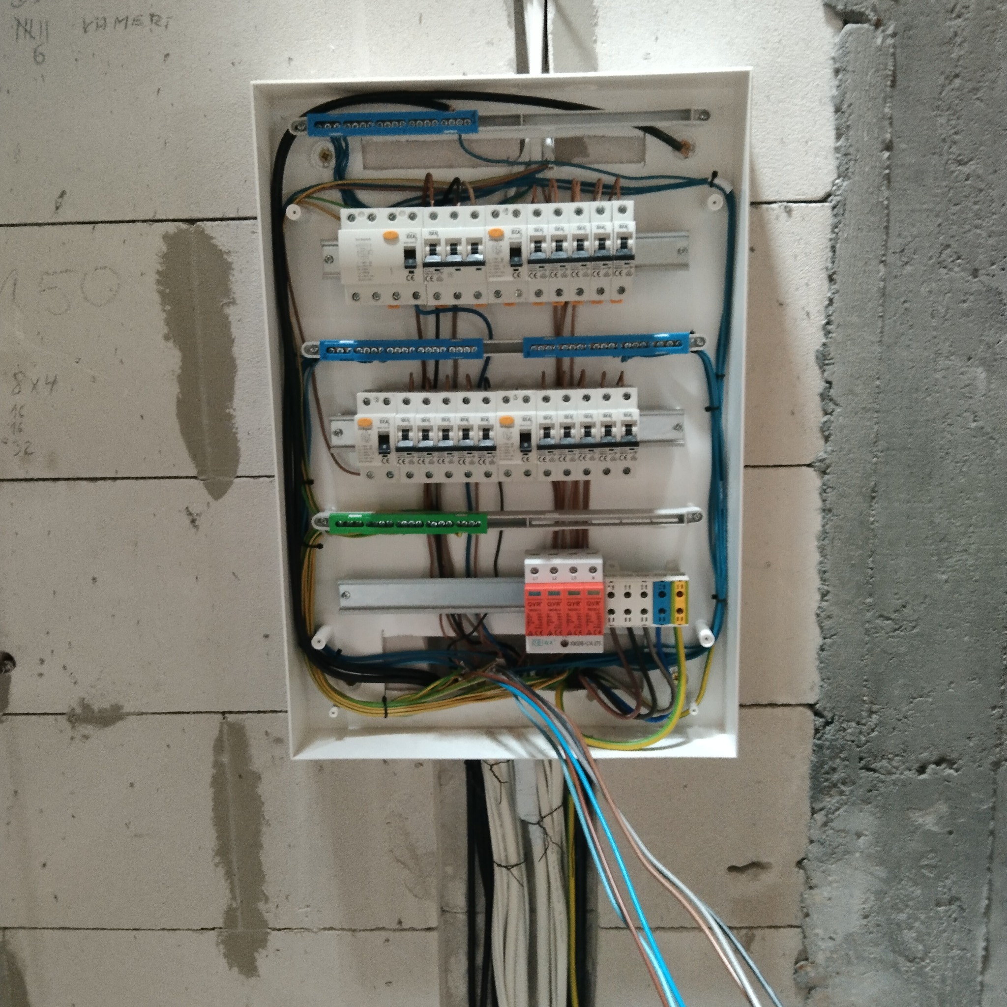 Otwarta skrzynka rozdzielcza z zabezpieczeniami, zamontowana na ścianie z betonu komórkowego, widoczne liczne przewody elektryczne w różnych kolorach.