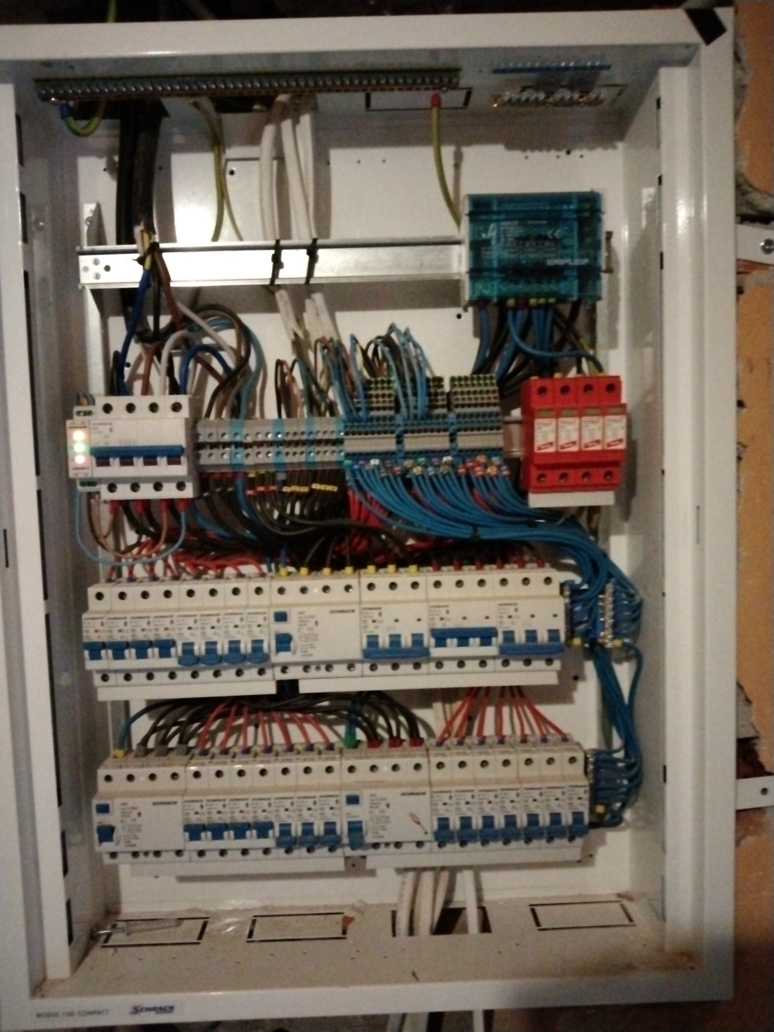 Rozdzielnica elektryczna w białej obudowie z widocznymi bezpiecznikami, przewodami w różnych kolorach, listwami zaciskowymi i ogranicznikiem przepięć, zamontowana przy ścianie.