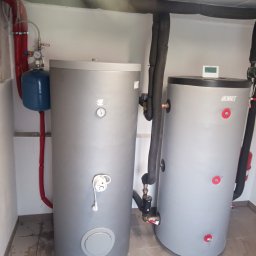 Firma Alan - Dwa szare zbiorniki akumulacyjne ciepła w kotłowni z widocznymi rurami w izolacji, manometrem i elementami instalacji hydraulicznej.