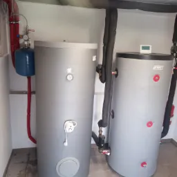 Dwa szare zbiorniki akumulacyjne ciepła w kotłowni z widocznymi rurami w izolacji, manometrem i elementami instalacji hydraulicznej.