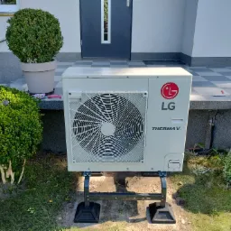 Zewnętrzna jednostka pompy ciepła LG Therma V, umieszczona na czarnej, metalowej konstrukcji, na tle wejścia do domu z szarą kostką brukową i zielenią.