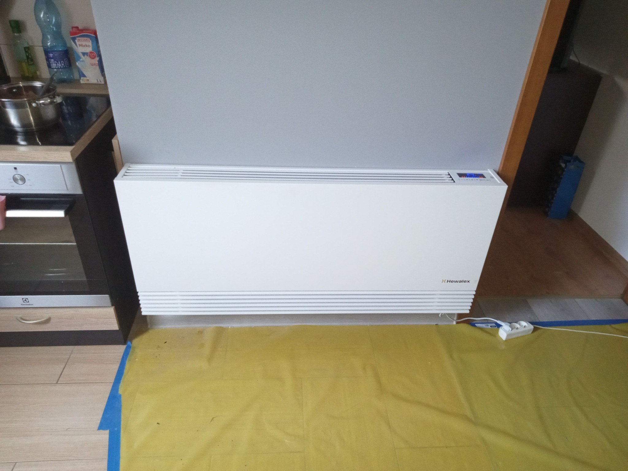 Biały grzejnik elektryczny Hewalex zamontowany w kuchni w Koziegłowach, zabezpieczony folią malarską na podłodze.