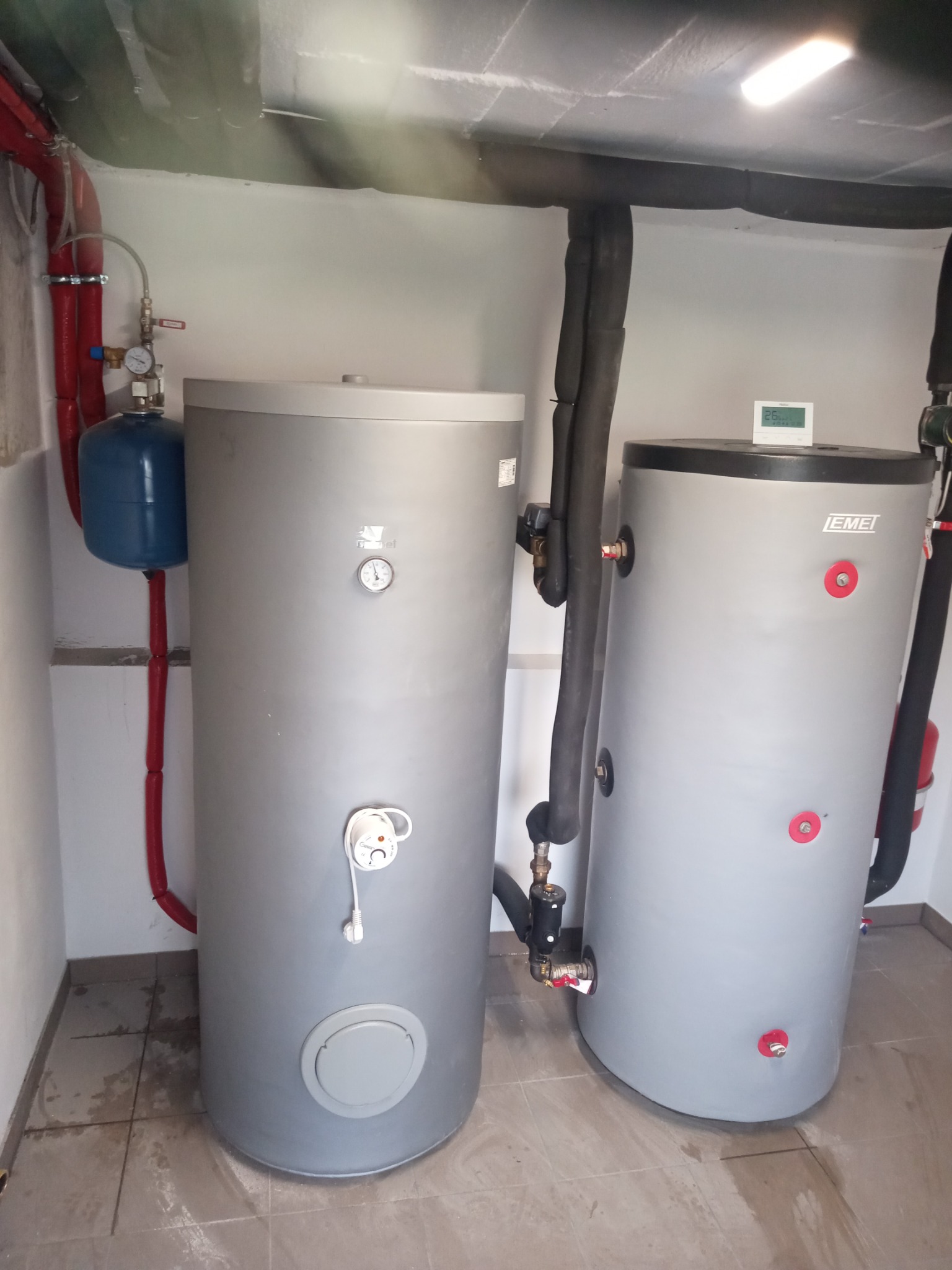 Dwa szare zbiorniki akumulacyjne ciepła w kotłowni z widocznymi rurami w izolacji, manometrem i elementami instalacji hydraulicznej.