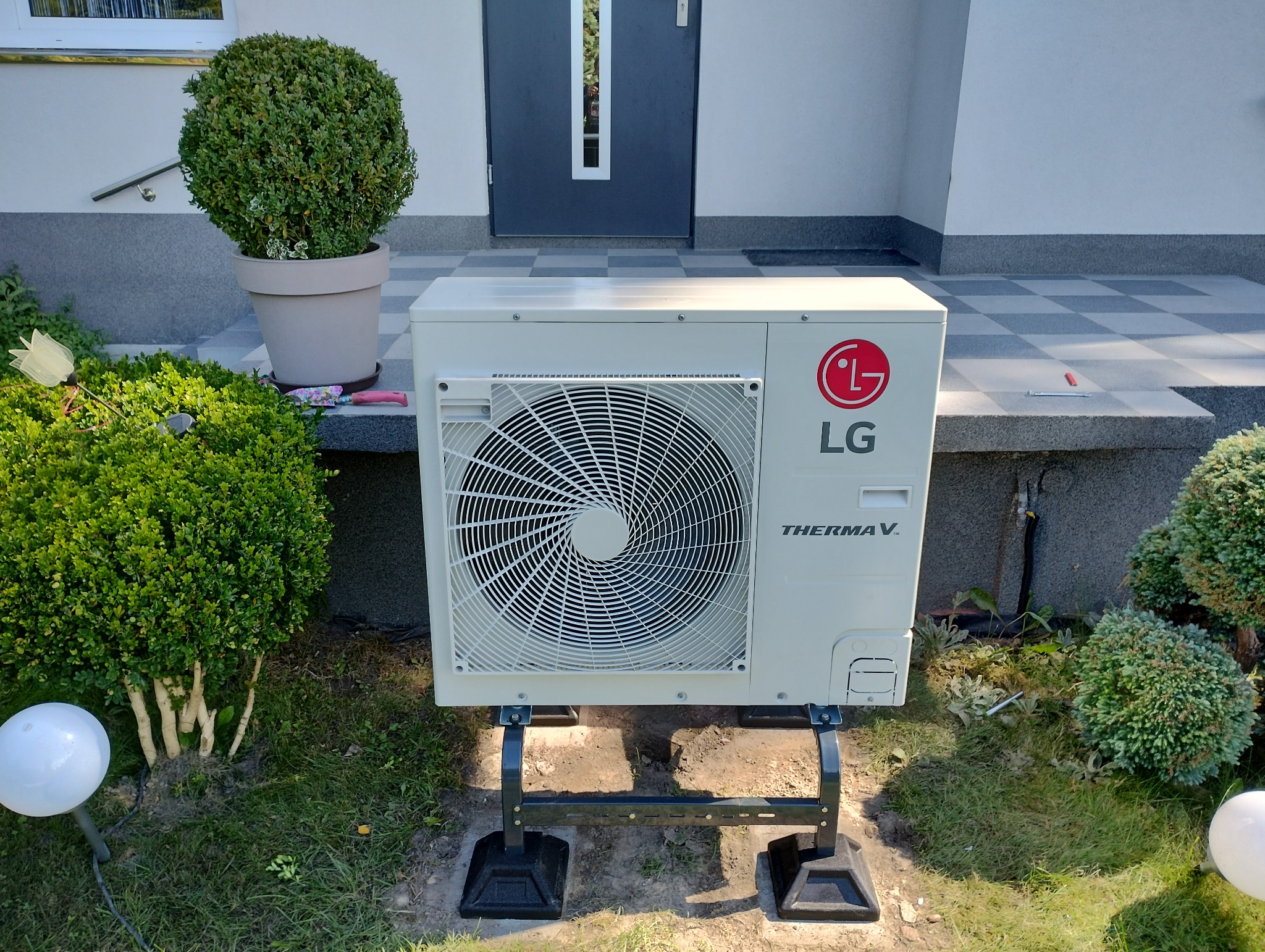 Zewnętrzna jednostka pompy ciepła LG Therma V, umieszczona na czarnej, metalowej konstrukcji, na tle wejścia do domu z szarą kostką brukową i zielenią.
