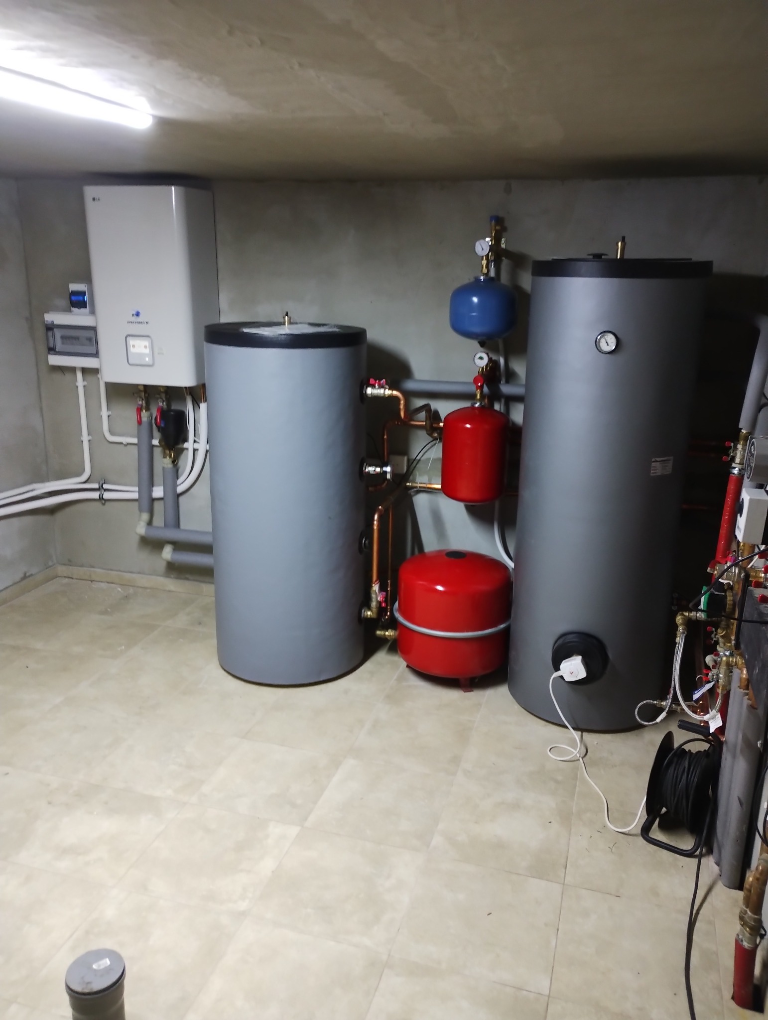 Kotłownia z dwoma szarymi zbiornikami akumulacyjnymi, czerwonymi naczyniami przeponowymi i białą pompą ciepła LG na ścianie, widoczne rury miedziane i białe plastikowe, na podłodze zwinięty kabel...