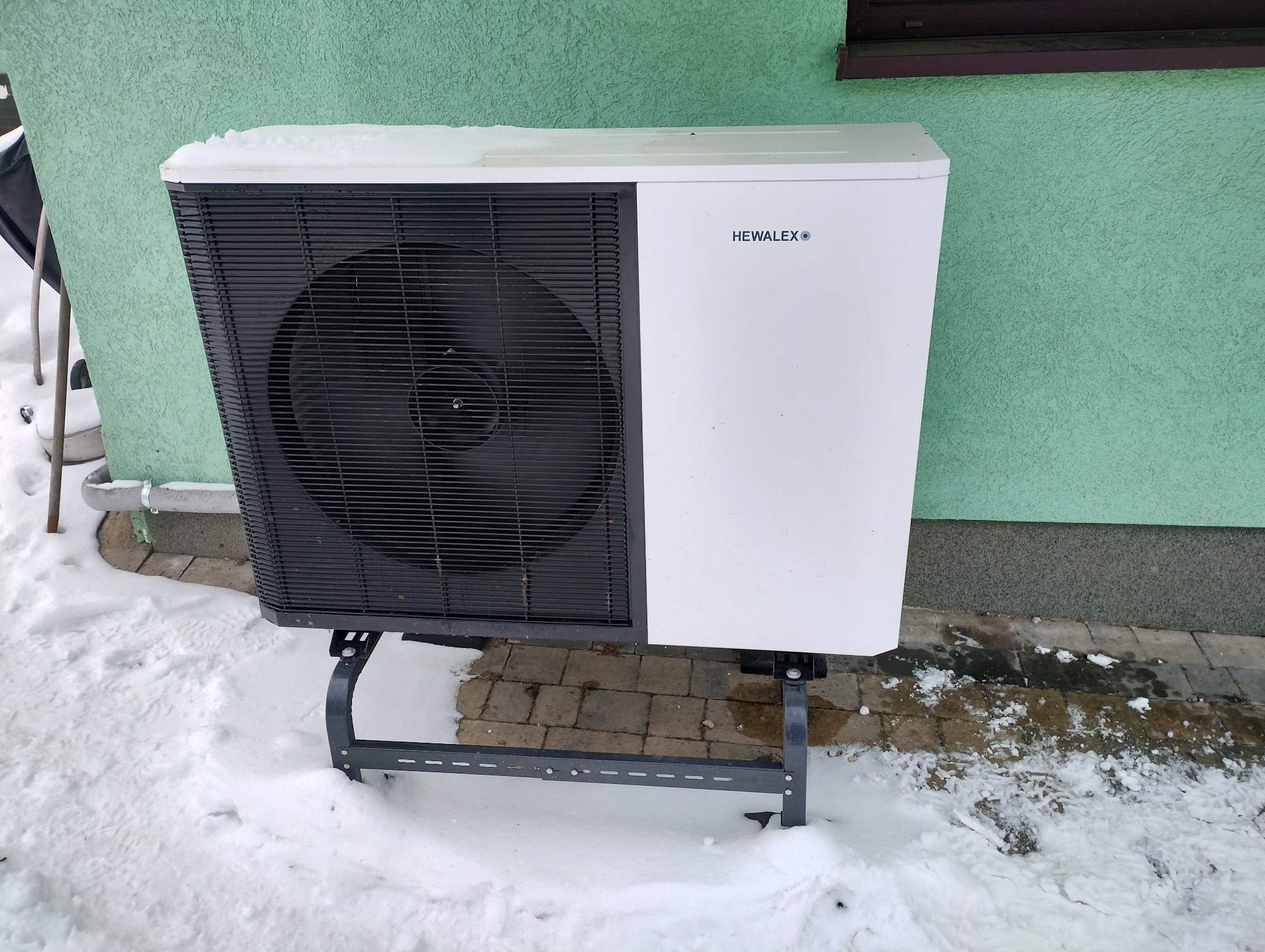 Biała jednostka zewnętrzna pompy ciepła marki Hewalex, zamontowana na metalowym stelażu, stoi na śniegu przy zielonej ścianie budynku.