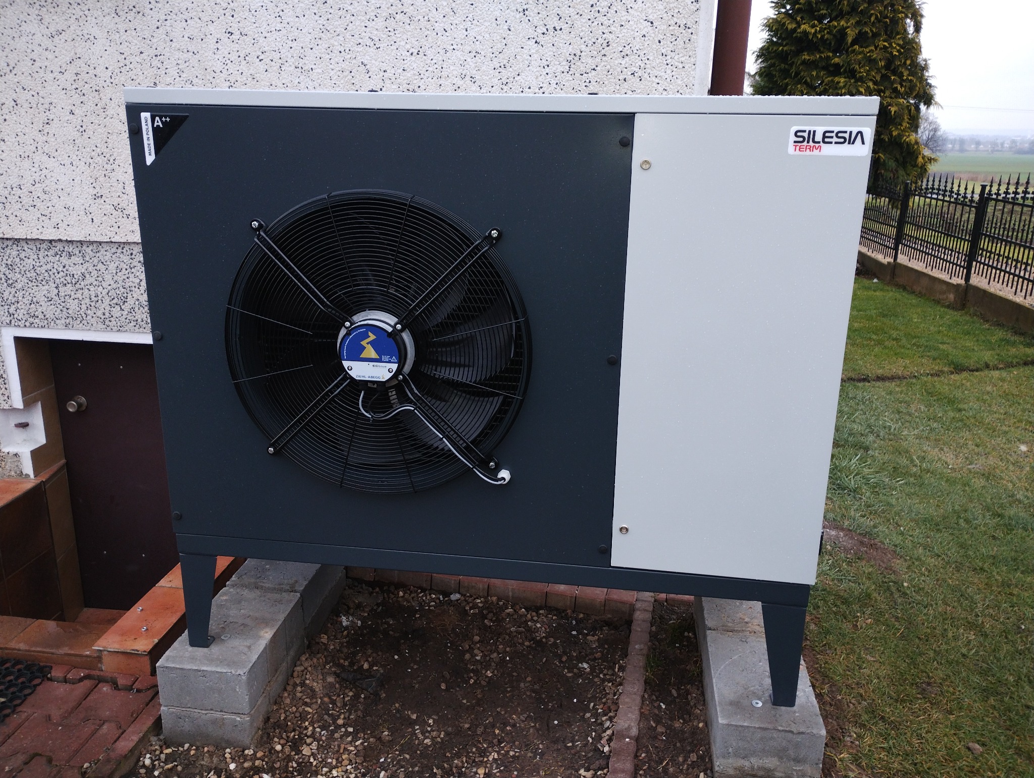 Zewnętrzna jednostka pompy ciepła Silesia Term, zamontowana na betonowych bloczkach przy ścianie budynku z widoczną etykietą A++ Made in Poland oraz wentylatorem z niebieskim logo, na tle trawnika...