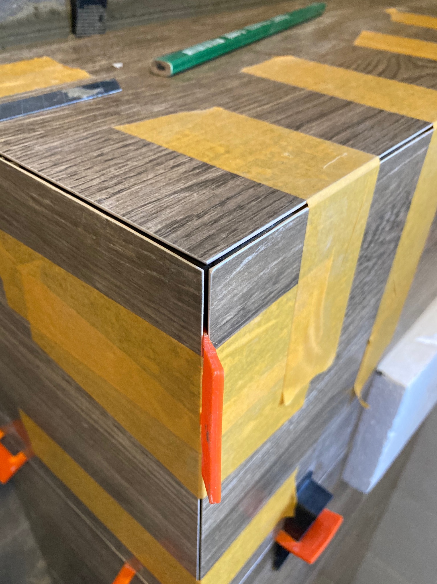 Fragment układania płytek imitujących drewno na krawędzi, z użyciem żółtej taśmy maskującej, pomarańczowych i czarnych klinów dystansowych oraz zielonego ołówka stolarskiego w tle.